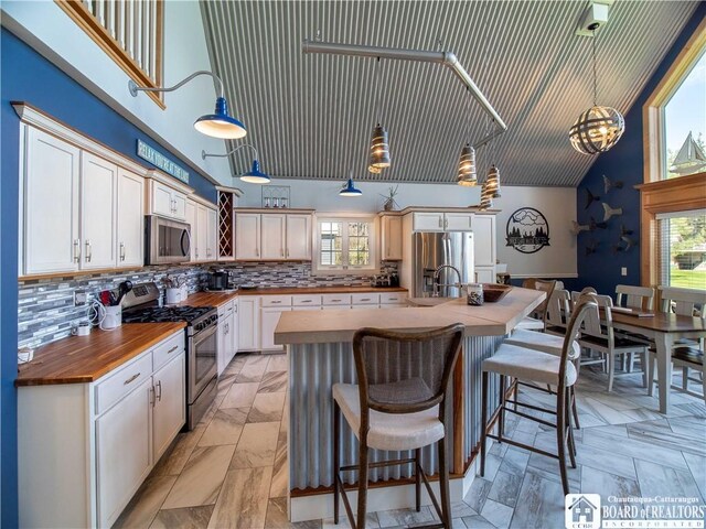kitchen featuring stainless steel appliances, a center island with sink, tasteful backsplash, a high ceiling, and butcher block counters