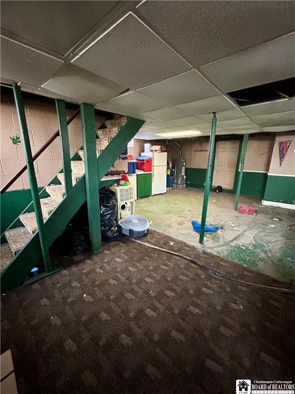 unfinished below grade area with a paneled ceiling, carpet flooring, freestanding refrigerator, water heater, and stairs