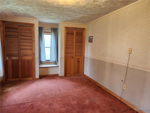 unfurnished bedroom featuring a textured ceiling, baseboards, carpet, wallpapered walls, and two closets