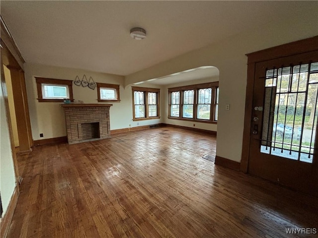 unfurnished living room featuring arched walkways, baseboards, a brick fireplace, and wood finished floors