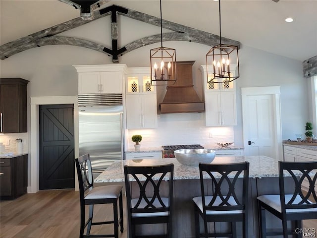 kitchen featuring an inviting chandelier, custom exhaust hood, stainless steel built in fridge, vaulted ceiling, and backsplash