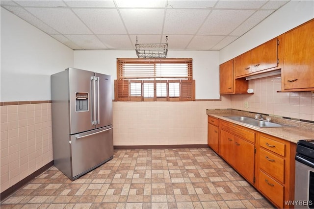 kitchen with a sink, stainless steel appliances, tile walls, a paneled ceiling, and brown cabinetry