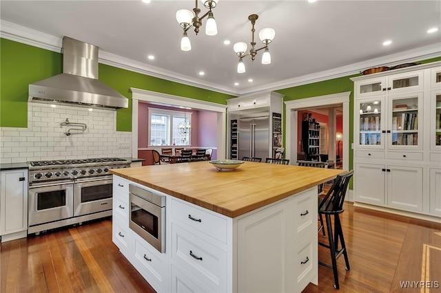 kitchen with wall chimney range hood, built in appliances, butcher block countertops, and crown molding