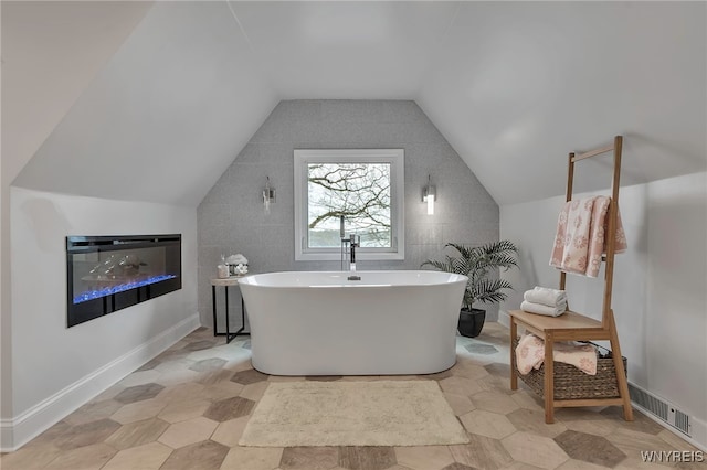full bath featuring a glass covered fireplace, visible vents, a freestanding bath, and vaulted ceiling