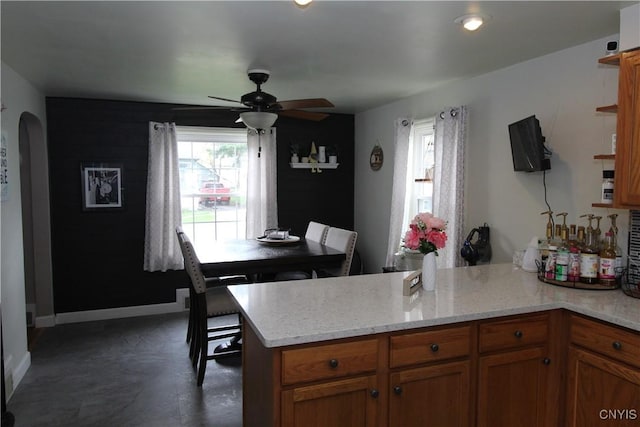 kitchen featuring a ceiling fan, arched walkways, light stone countertops, brown cabinets, and a peninsula
