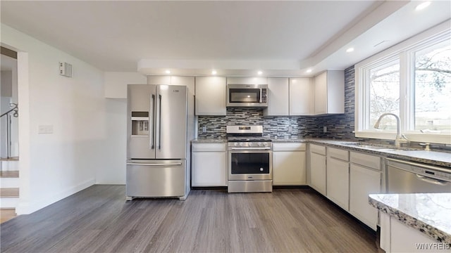 kitchen with a sink, appliances with stainless steel finishes, light stone countertops, wood finished floors, and backsplash