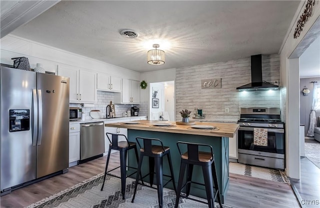kitchen featuring wood finished floors, butcher block countertops, appliances with stainless steel finishes, white cabinetry, and wall chimney exhaust hood