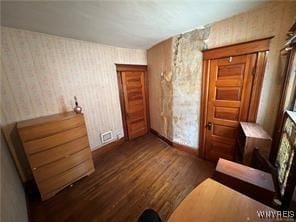 unfurnished bedroom with visible vents, wallpapered walls, dark wood finished floors, and baseboards