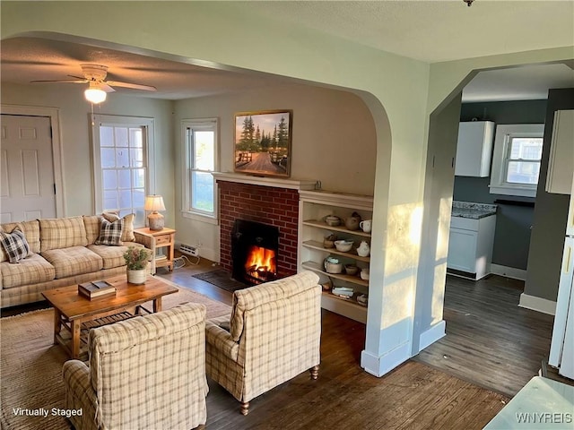 living area with arched walkways, plenty of natural light, a brick fireplace, dark wood-style floors, and a ceiling fan