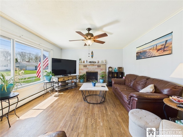 living room with light wood-style floors, baseboard heating, ornamental molding, a fireplace, and a ceiling fan