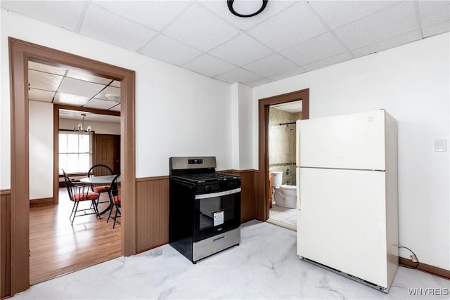 kitchen featuring stainless steel range with gas stovetop, freestanding refrigerator, wainscoting, and a drop ceiling