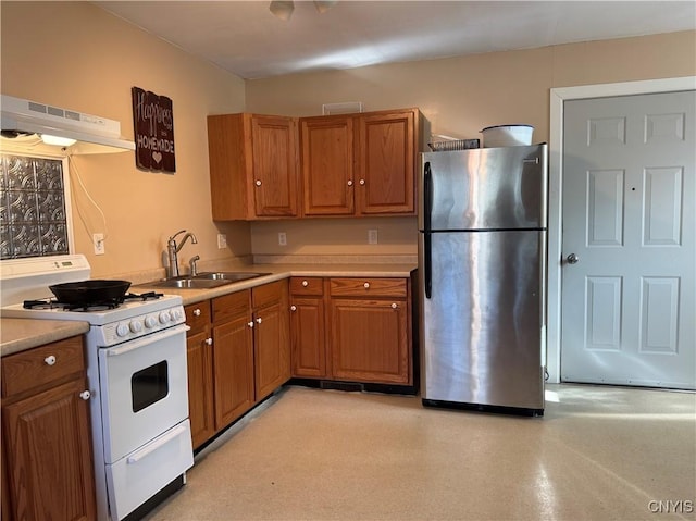 kitchen featuring freestanding refrigerator, under cabinet range hood, brown cabinets, white range with gas cooktop, and a sink
