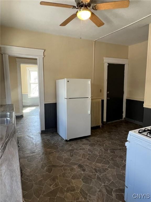 kitchen featuring stone flooring, ceiling fan, a wainscoted wall, radiator heating unit, and white appliances