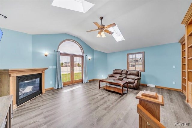 living area featuring light wood-style floors, a glass covered fireplace, lofted ceiling with skylight, a ceiling fan, and baseboards