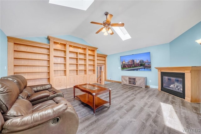 living room featuring wood finished floors, vaulted ceiling with skylight, a glass covered fireplace, baseboards, and a ceiling fan