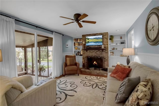 living room featuring built in shelves, a ceiling fan, a brick fireplace, and wainscoting