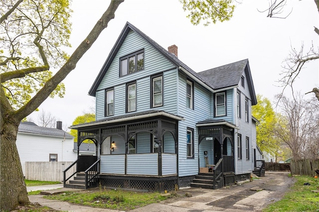 victorian-style house featuring roof with shingles, fence, covered porch, and a chimney