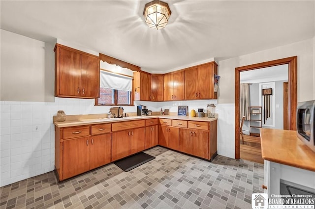kitchen with brown cabinetry, tile walls, light countertops, a wainscoted wall, and a sink