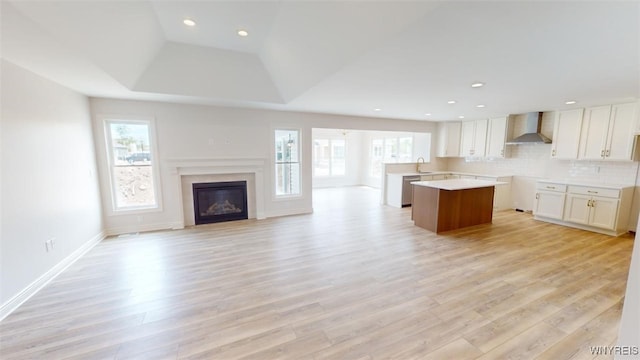 kitchen featuring light wood finished floors, a glass covered fireplace, a tray ceiling, wall chimney range hood, and open floor plan
