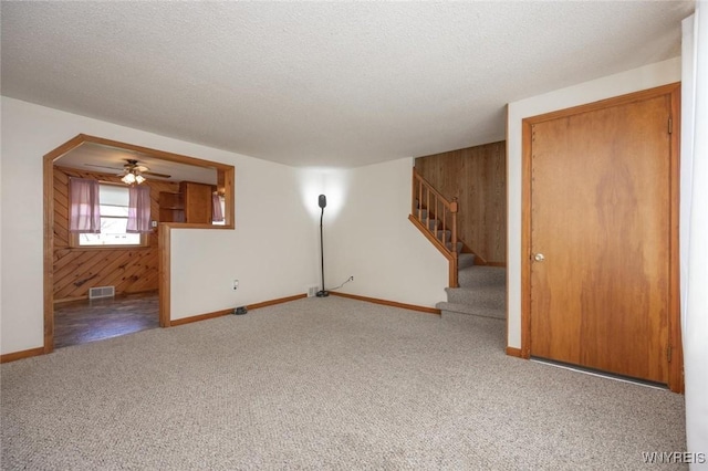 unfurnished living room with carpet flooring, visible vents, baseboards, wooden walls, and stairs