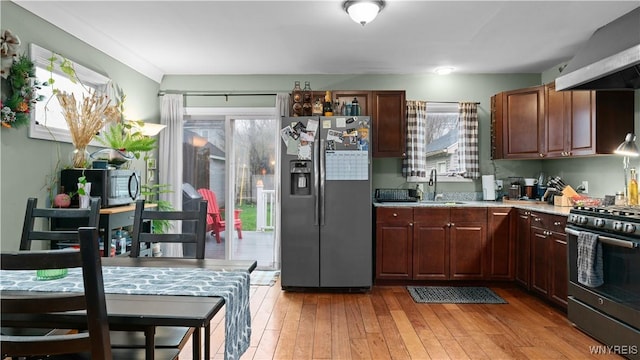 kitchen with light countertops, wall chimney exhaust hood, a sink, light wood-style flooring, and appliances with stainless steel finishes