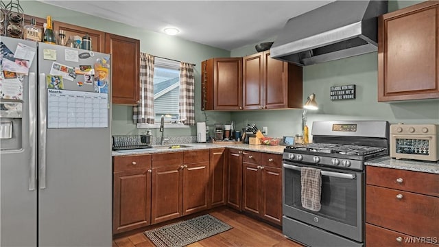 kitchen featuring wall chimney exhaust hood, light stone counters, a sink, light wood-style flooring, and appliances with stainless steel finishes