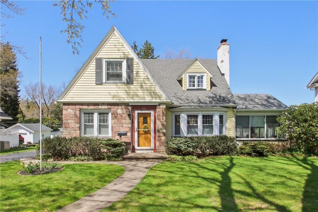 cape cod home featuring a chimney, roof with shingles, a front yard, and stone siding