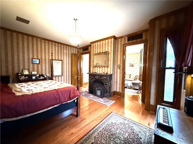bedroom featuring visible vents, crown molding, wood finished floors, baseboards, and wallpapered walls