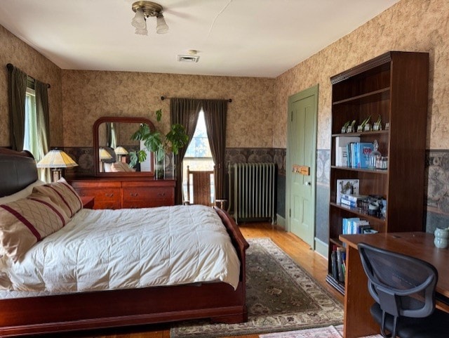 bedroom featuring radiator heating unit, a wainscoted wall, wallpapered walls, wood finished floors, and visible vents