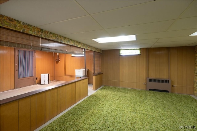basement featuring radiator heating unit, a drop ceiling, wooden walls, and light colored carpet