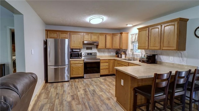 kitchen with a peninsula, light wood-type flooring, under cabinet range hood, a sink, and appliances with stainless steel finishes