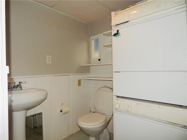bathroom featuring a sink, wainscoting, stacked washing maching and dryer, and toilet