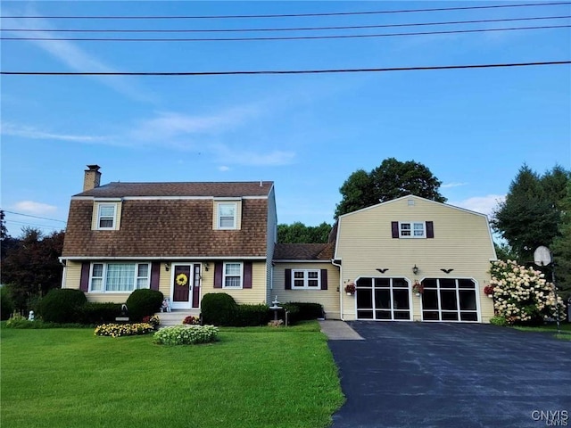 colonial inspired home with driveway, a chimney, a front yard, a gambrel roof, and a shingled roof