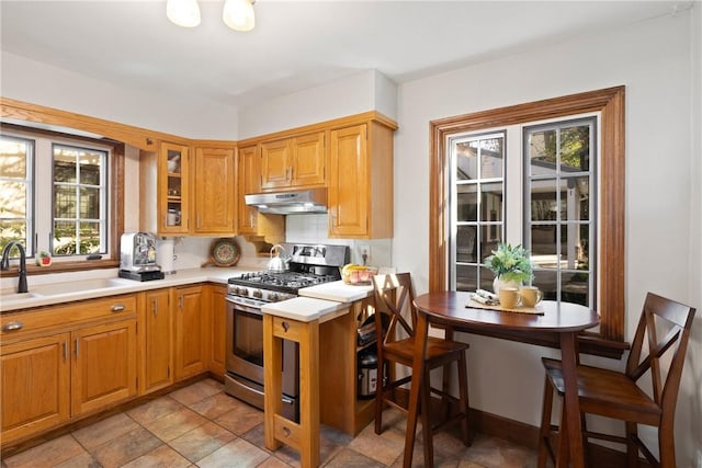 kitchen featuring glass insert cabinets, stainless steel gas range oven, light countertops, under cabinet range hood, and a sink