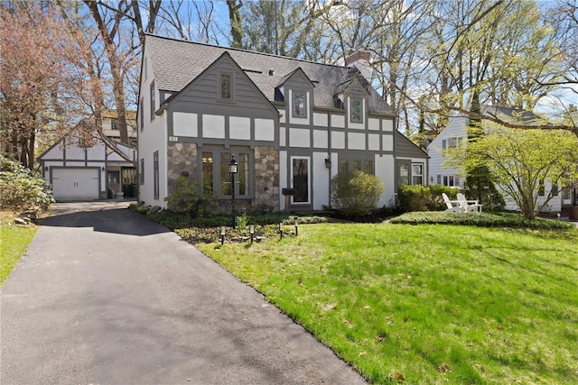 tudor house featuring aphalt driveway, a chimney, an outdoor structure, a front lawn, and stucco siding