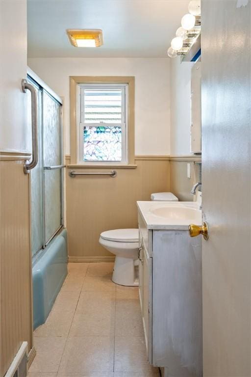 full bathroom featuring vanity, tile patterned flooring, wainscoting, toilet, and bath / shower combo with glass door