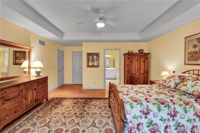 bedroom with light wood finished floors, ensuite bath, a ceiling fan, a tray ceiling, and visible vents