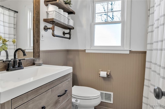 full bath featuring visible vents, vanity, curtained shower, toilet, and a wainscoted wall