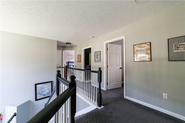 hall featuring a textured ceiling, baseboards, an upstairs landing, and dark colored carpet