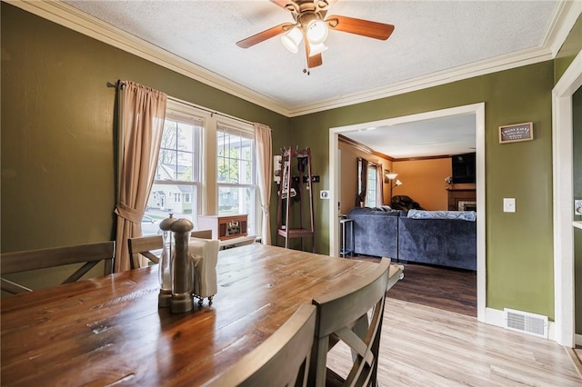 dining room with crown molding, ceiling fan, a fireplace, light wood-style flooring, and visible vents