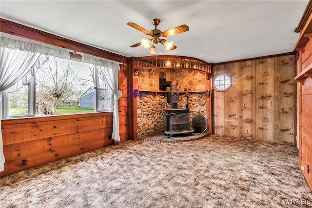 unfurnished living room with a ceiling fan, a wood stove, wood walls, and carpet flooring