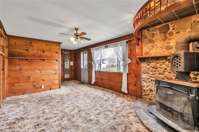 unfurnished living room featuring carpet floors, a wood stove, ceiling fan, and wooden walls