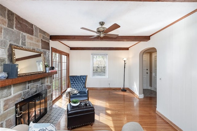 living area with beam ceiling, a fireplace, arched walkways, a ceiling fan, and wood finished floors