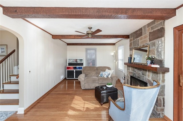 living room featuring stairs, beam ceiling, wood finished floors, a stone fireplace, and ceiling fan