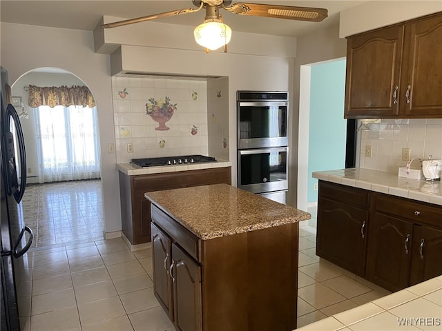 kitchen with freestanding refrigerator, gas cooktop, stainless steel double oven, tile countertops, and arched walkways