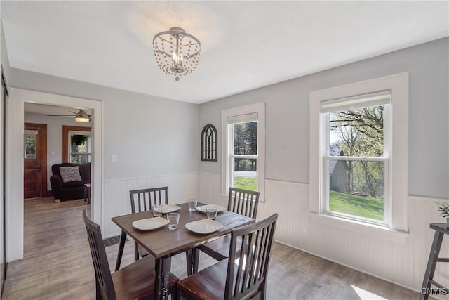 dining area with ceiling fan with notable chandelier, plenty of natural light, wainscoting, and wood finished floors