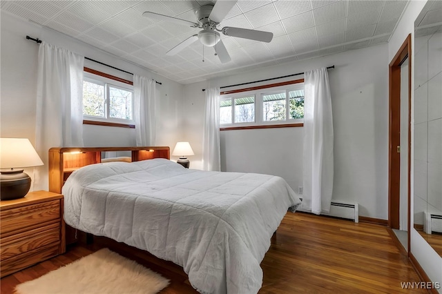 bedroom featuring ceiling fan, multiple windows, a baseboard heating unit, and wood finished floors
