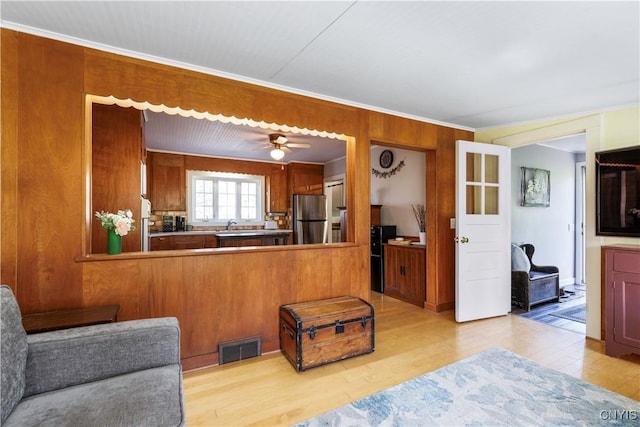 living room featuring wooden walls, ceiling fan, light wood-style flooring, visible vents, and ornamental molding