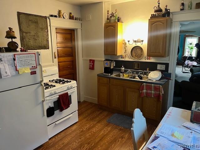 kitchen with dark wood-style flooring, dark countertops, white appliances, brown cabinets, and a sink