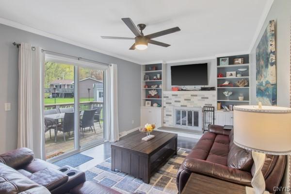 living area with ornamental molding, a glass covered fireplace, baseboards, ceiling fan, and built in shelves